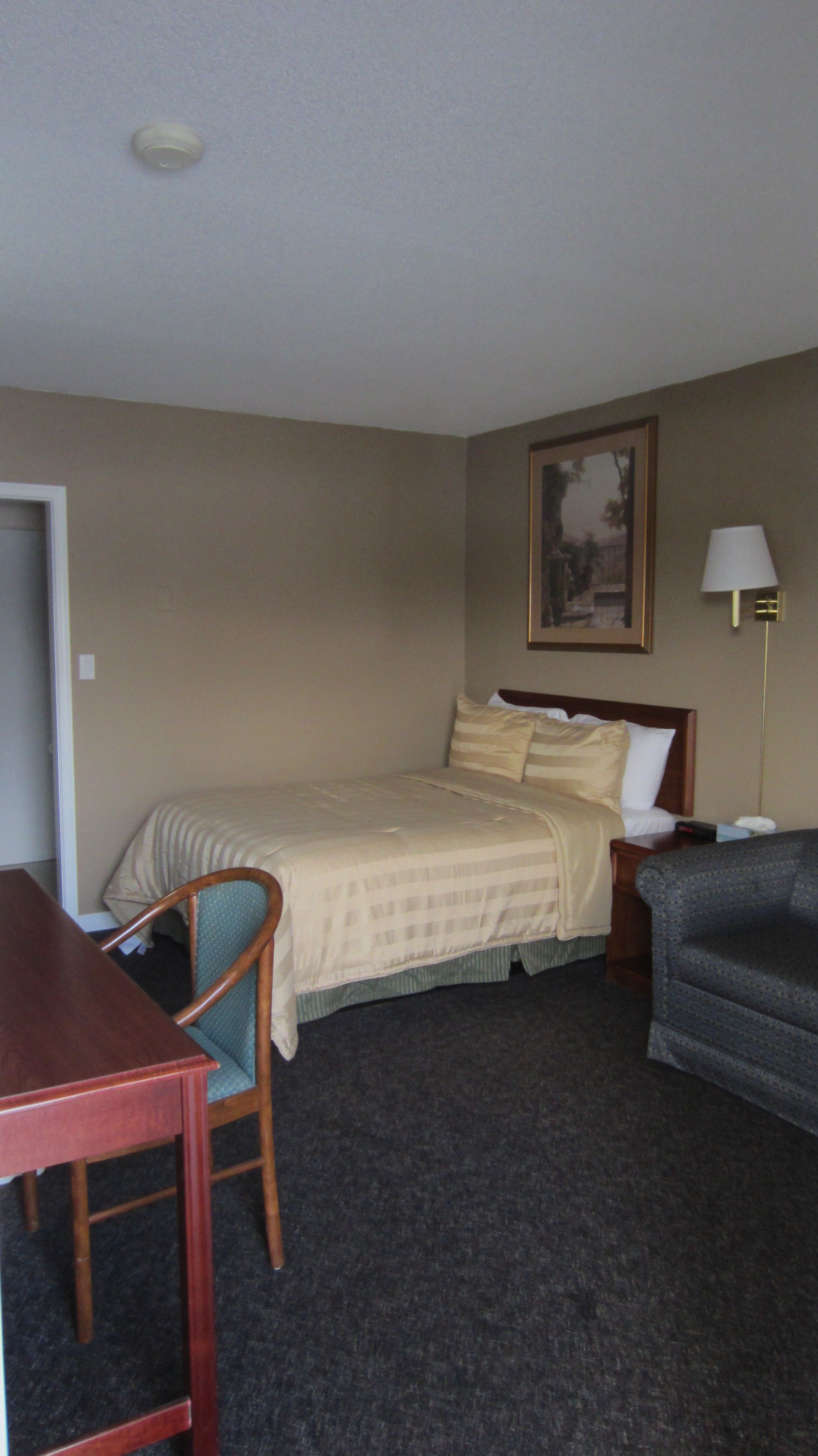 Una habitación de hotel con una cama, una silla, un escritorio, una lámpara y un cuadro en la pared.
