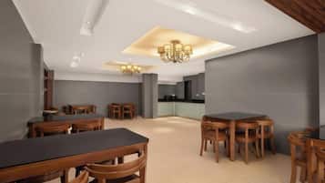 A modern dining area with multiple wooden tables and chairs, a kitchen in the background, and recessed lighting on the ceiling.