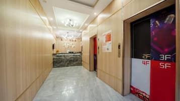 Un pasillo de hotel con mostrador de recepción, puertas de ascensor indicando desde el 2F hasta el 5F y un candelabro en el techo.