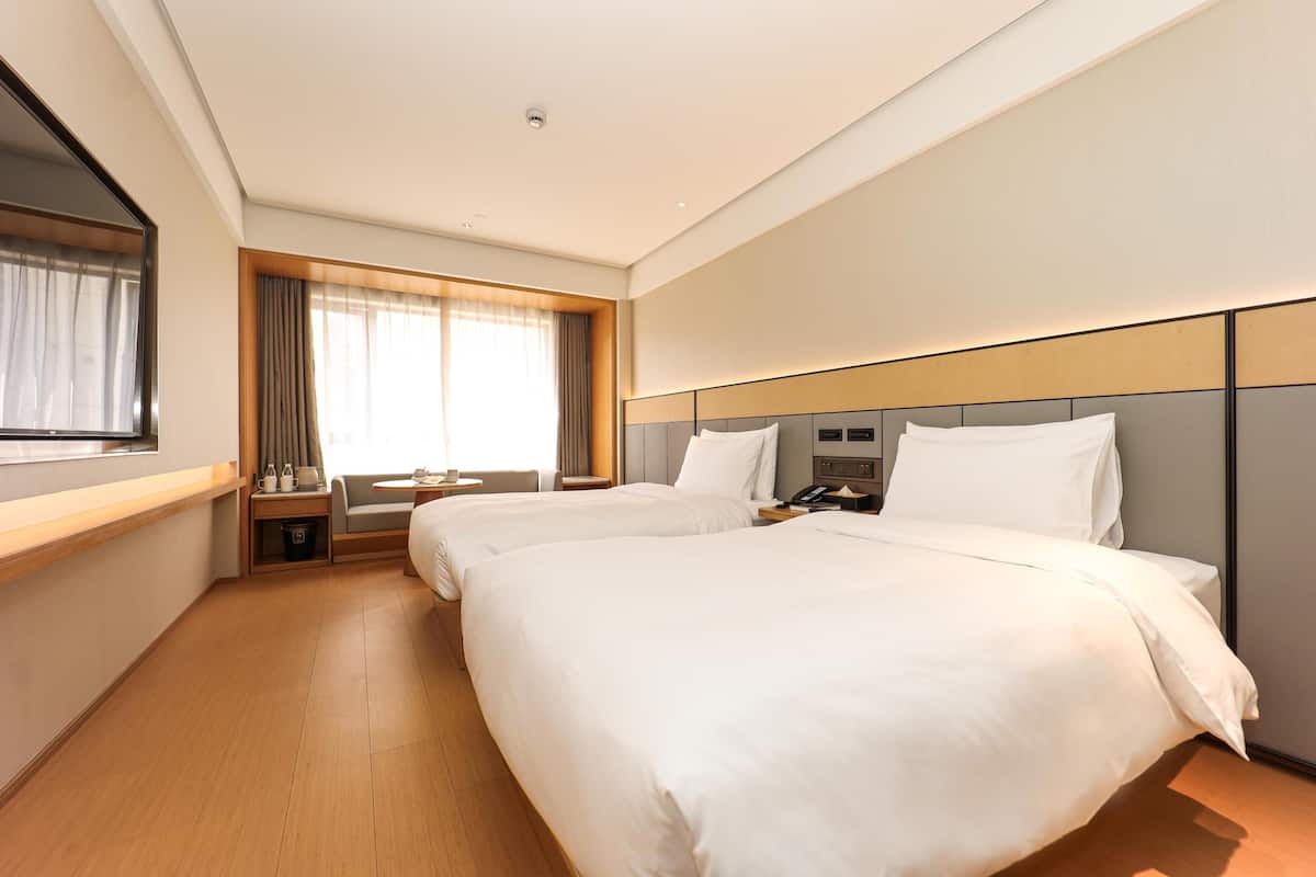 Quarto de hotel com duas camas, uma escrivaninha, uma cadeira, uma janela com cortinas e uma mesinha de cabeceira.