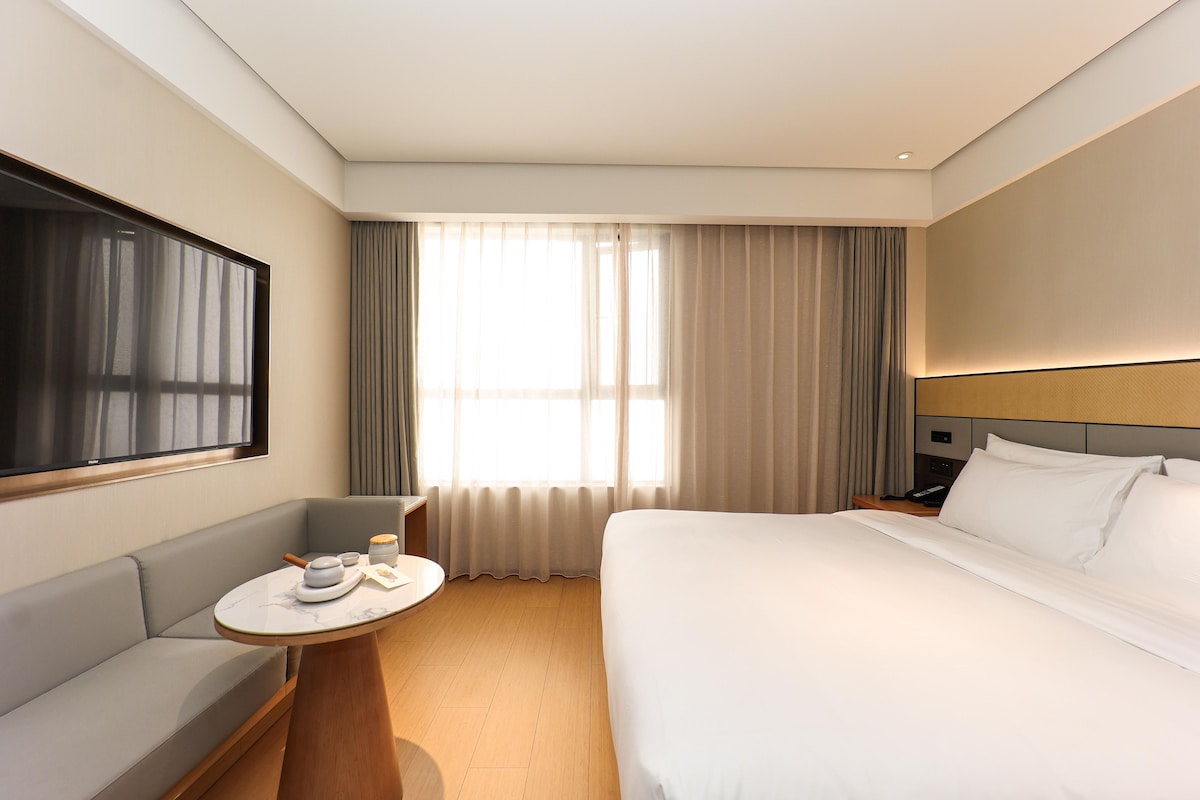 A hotel room with a bed, a sofa, a small table with a remote control, and a window with curtains.