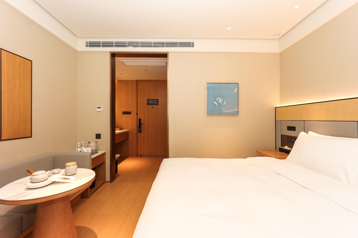 A hotel room with a bed, a bedside table, a small round table with kitchenware, a shower area, and a wooden door.