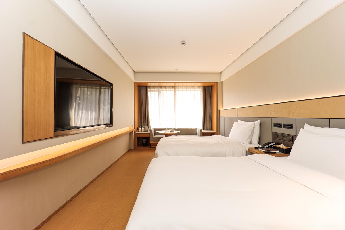 Habitació d'hotel amb dos llits, televisió, escriptori i finestra amb cortines.
