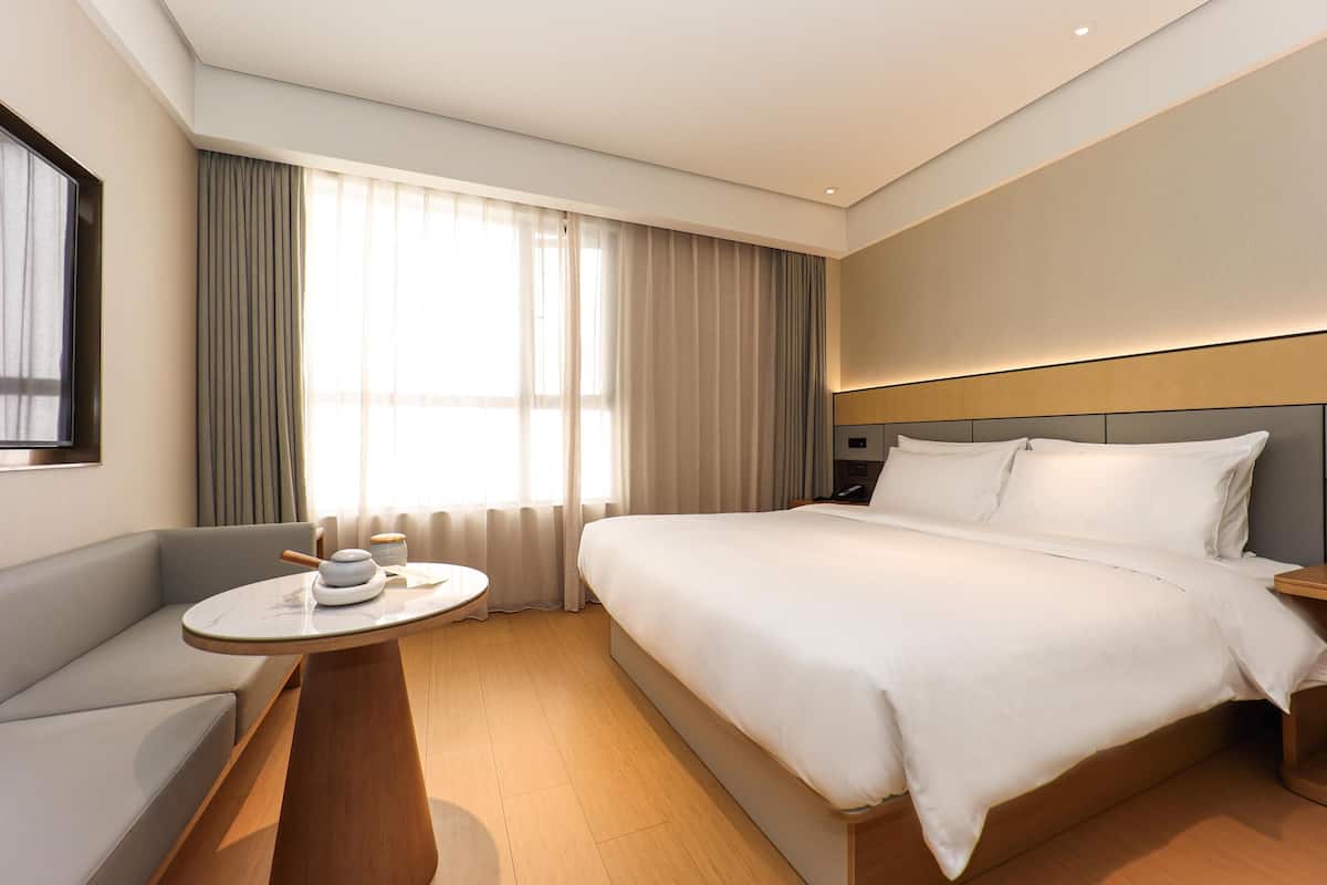 Quarto de hotel com cama, sofá, mesa redonda com cafeteira e uma janela grande com cortinas.