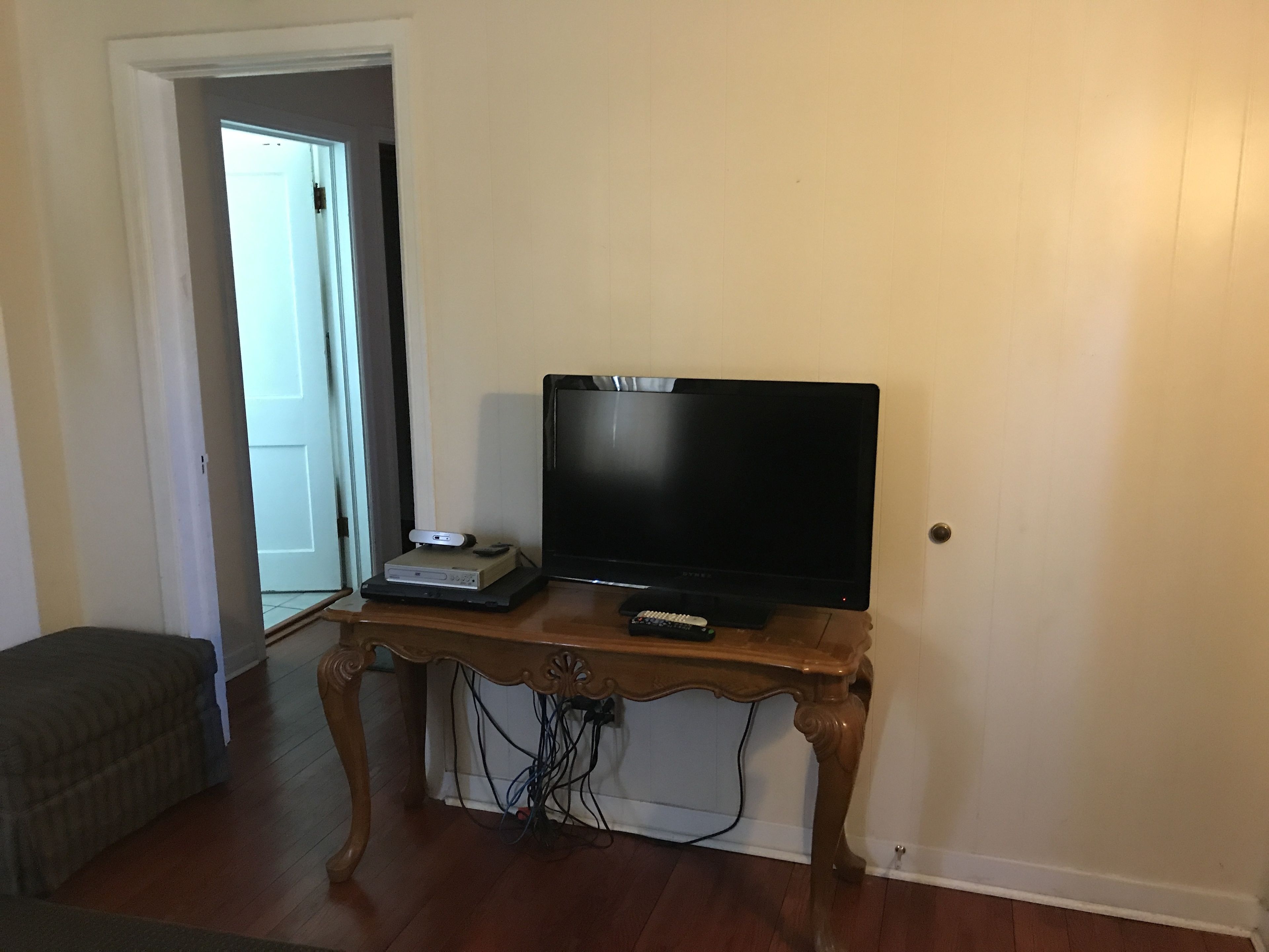 A living room with a wooden table holding a flat-screen TV, a DVD player, and a remote control. There is a couch in the foreground and a door leading to another room.