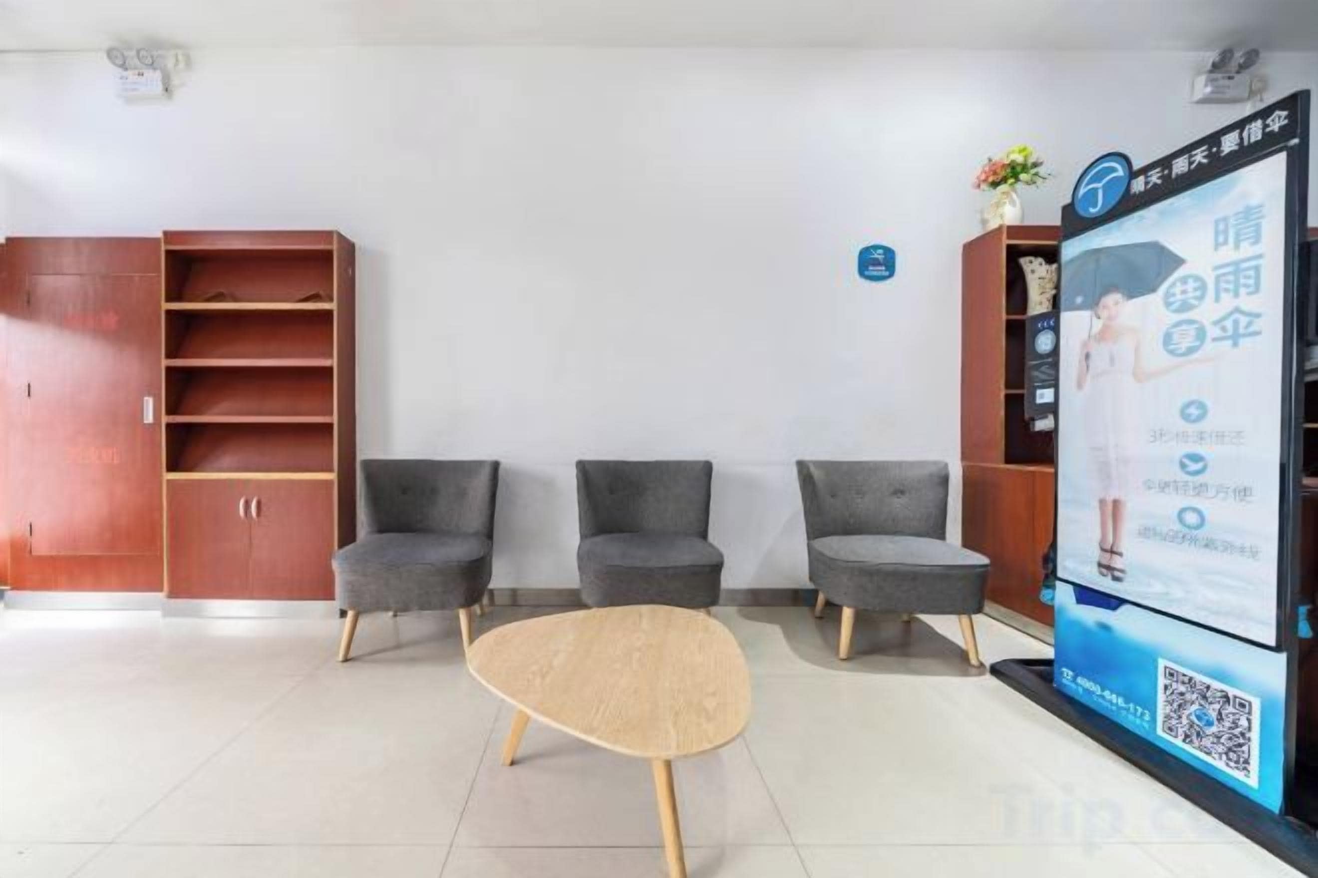 A waiting area with three grey chairs, a wooden table, and a digital screen displaying information.