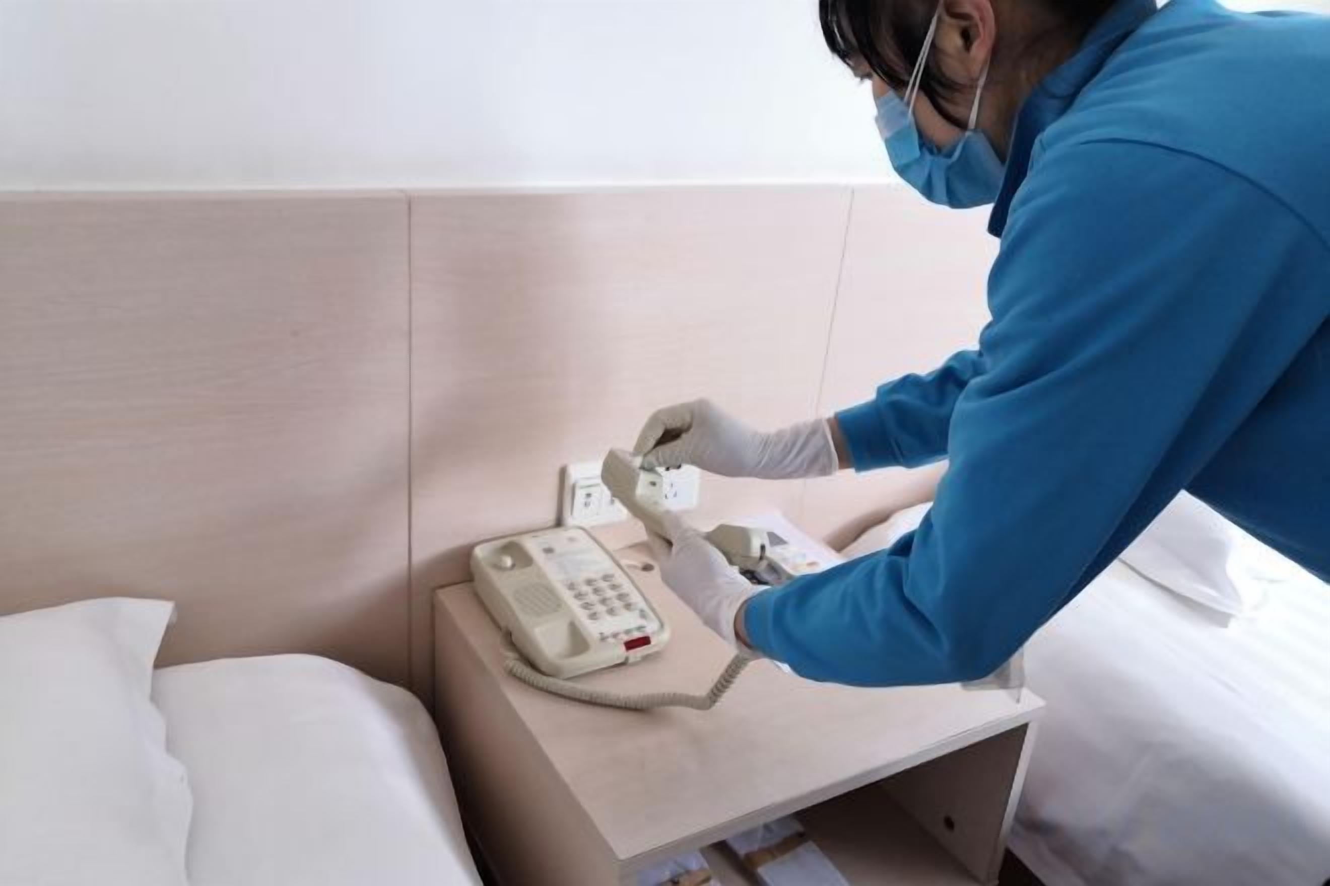 A person in a blue uniform and gloves handling a telephone on a bedside table in a hotel room.