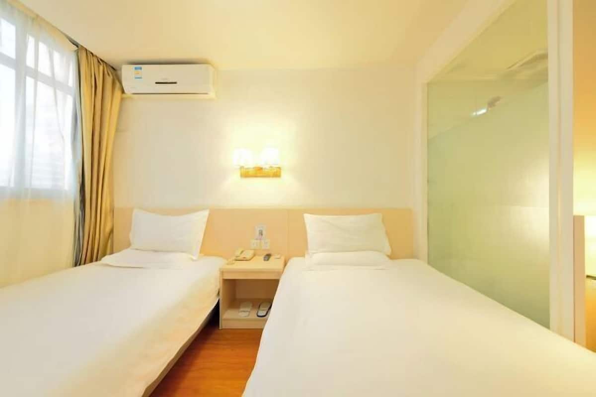 Une chambre d’hôtel avec deux lits, une table de chevet avec un téléphone et une lampe, un appareil de climatisation et une fenêtre avec des rideaux.