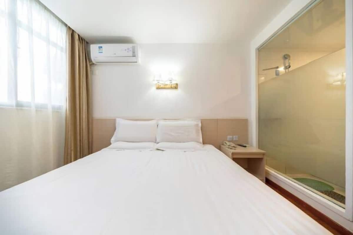 Une chambre d’hôtel comprenant un lit, une table de chevet, un climatiseur fixé au mur et une porte-fenêtre coulissante donnant sur un espace douche.