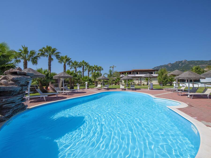 Piscina con sillones de descanso y palmeras, rodeada por un edificio con montañas al fondo.