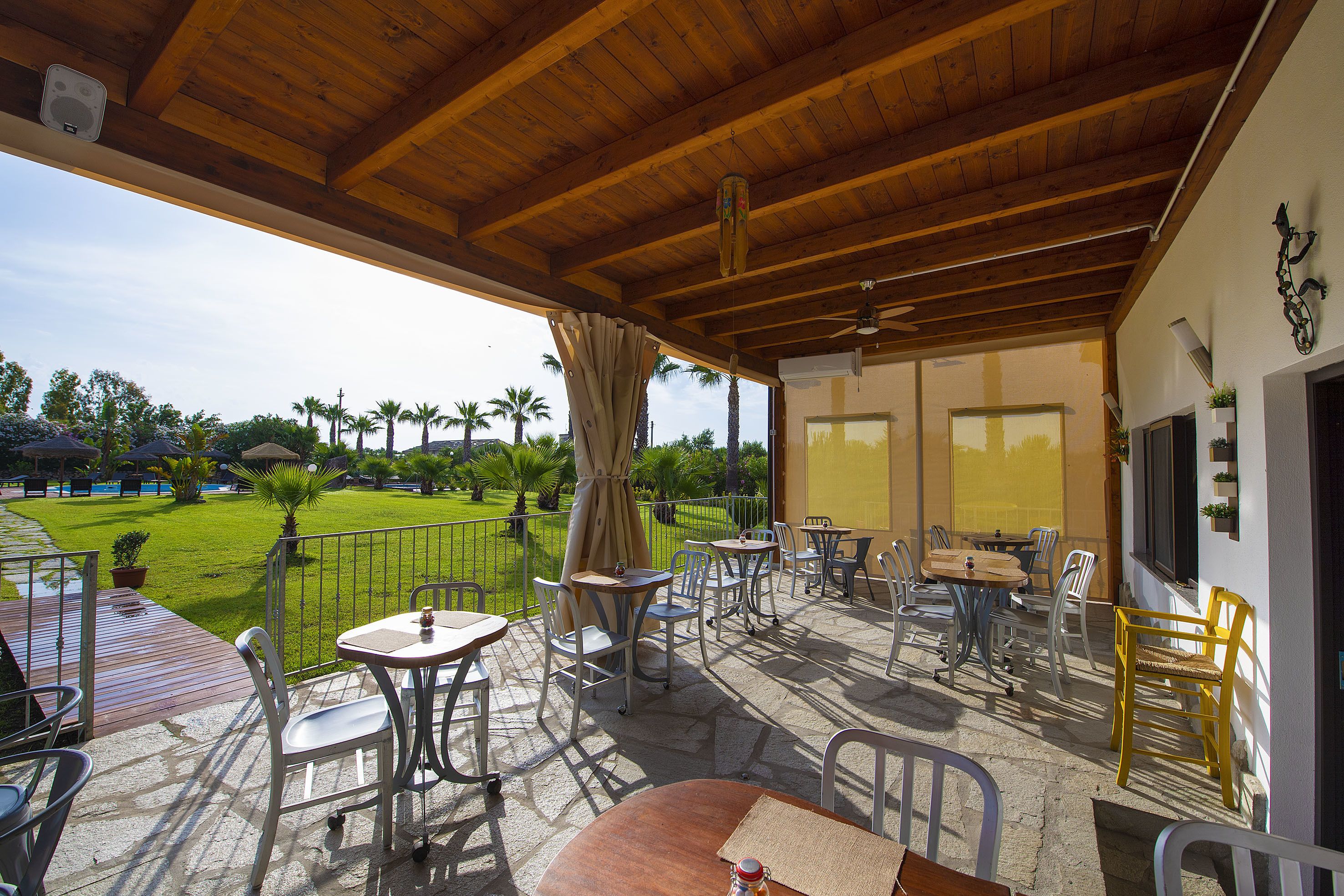 A covered outdoor patio with tables and chairs, a wooden ceiling, and a view of a green lawn and palm trees.