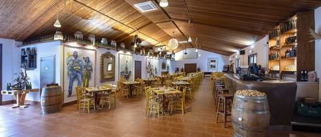A spacious restaurant with wooden ceilings and tiled floors, featuring multiple dining tables and chairs, a bar area, and wine racks.