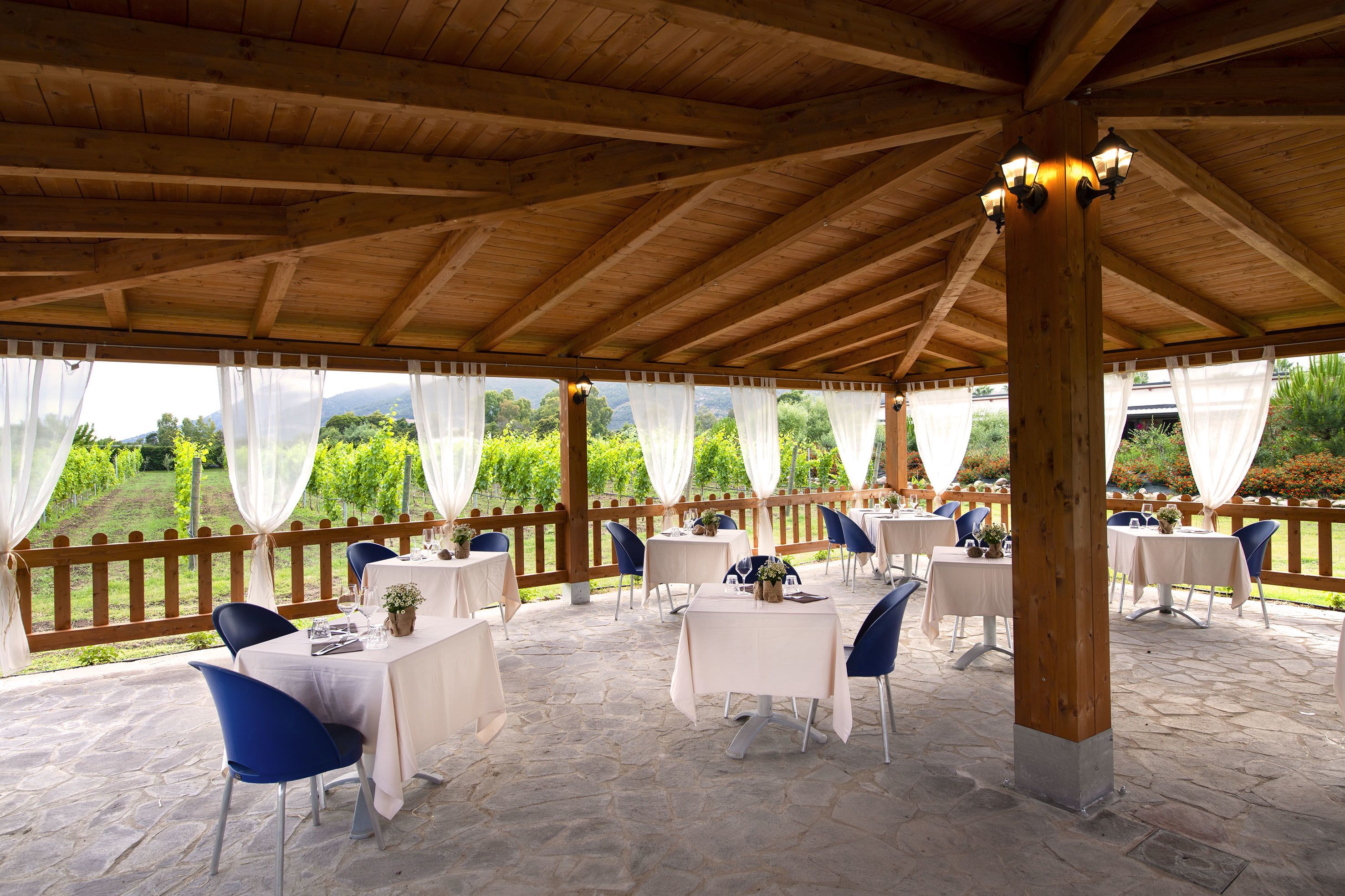 Une terrasse extérieure couverte, dotée d’un sol en pierre, de poutres en bois, de nappes blanches et de chaises bleues.