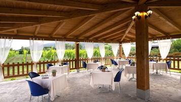 Une terrasse extérieure couverte, dotée d’un sol en pierre, de poutres en bois, de nappes blanches et de chaises bleues.