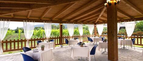 Eine überdachte Terrasse im Freien mit Steinboden, Holzbalken, weißen Tischdecken und blauen Stühlen.