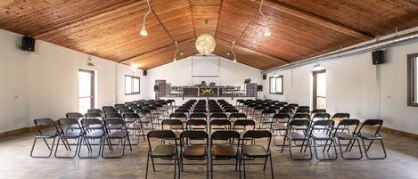 Un amplio salón con filas de sillas dispuestas para un evento, techo de madera y luz natural que entra por las ventanas.