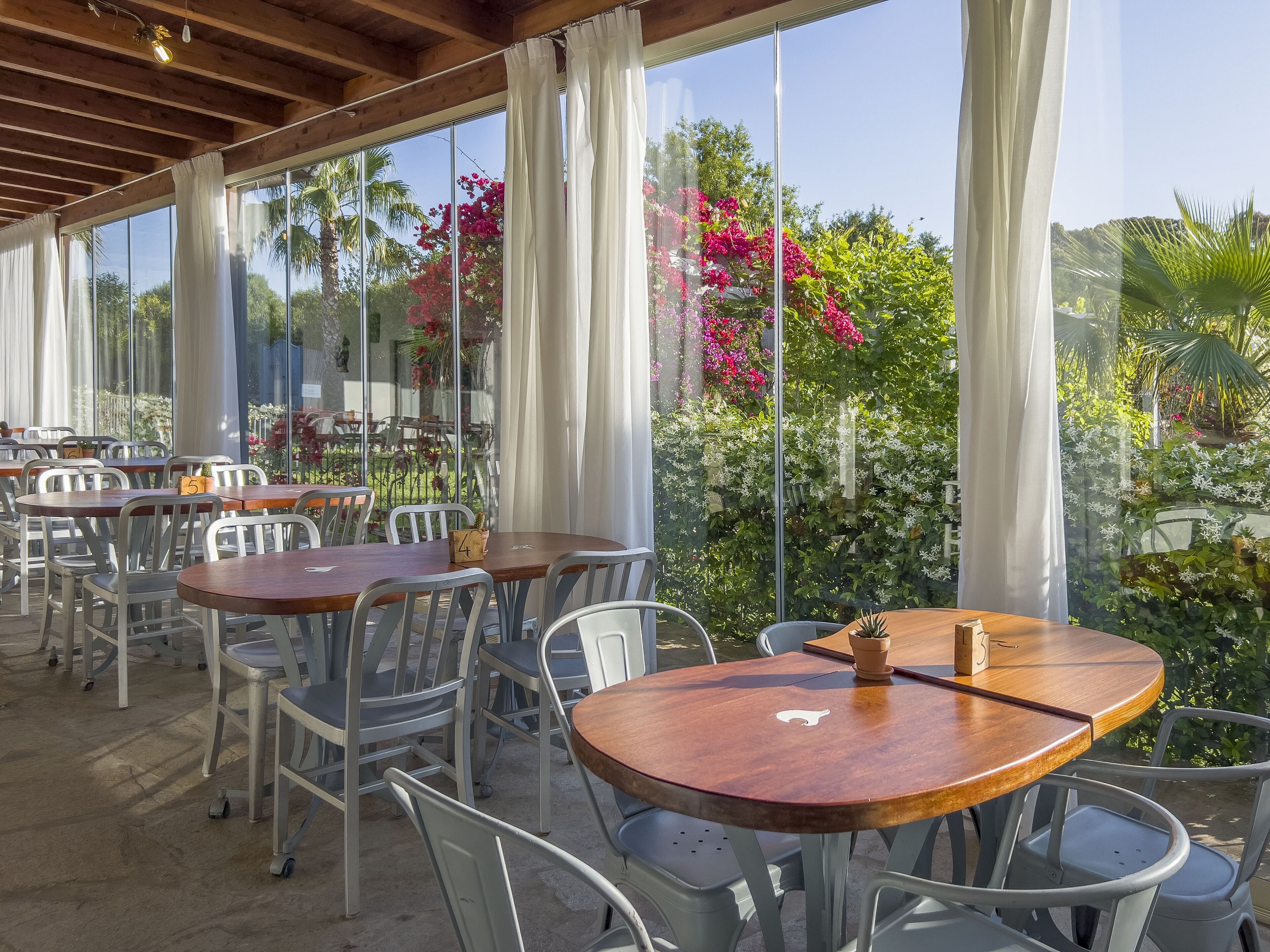 Un área de comedor al aire libre con mesas y sillas, ventanas amplias y vistas a follaje y flores.