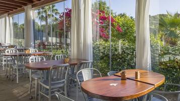 Un área de comedor al aire libre con mesas y sillas, ventanas amplias y vistas a follaje y flores.
