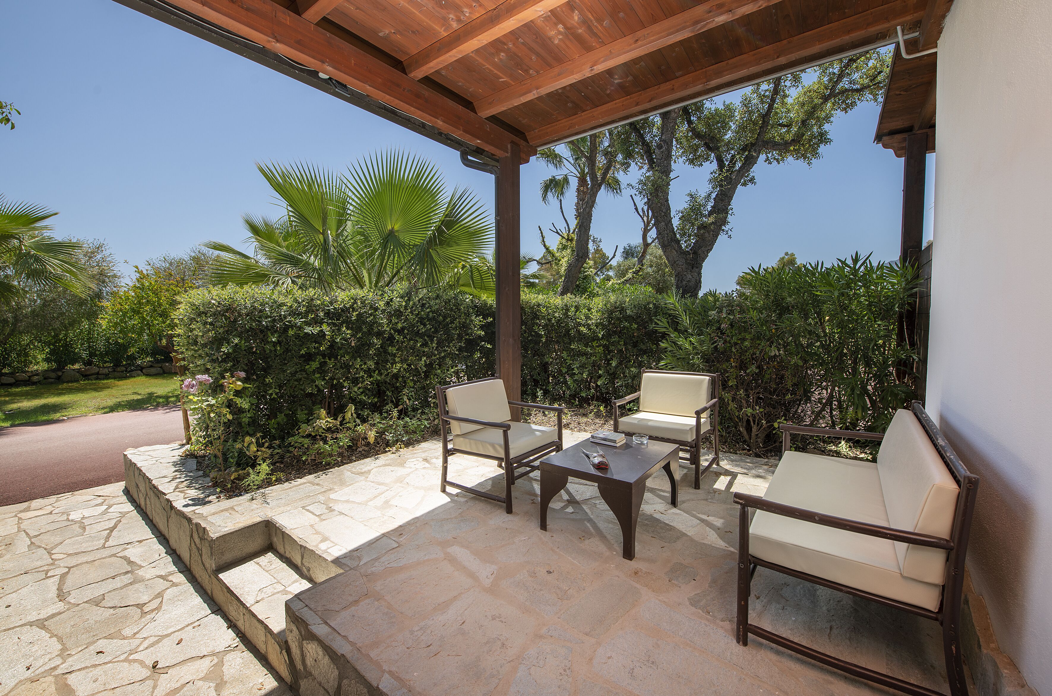 Eine Terrasse im Freien mit Steinboden, zwei beigefarbenen Polsterstühlen, einem Holztisch und einer Pergola darüber.