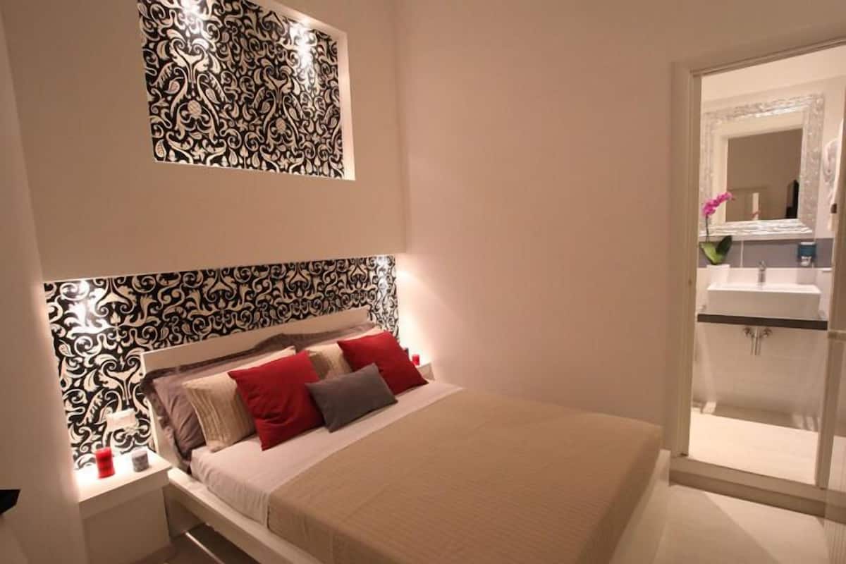 A modern bedroom with a patterned headboard, a bed with red and gray pillows, and a view of a bathroom through a glass door.