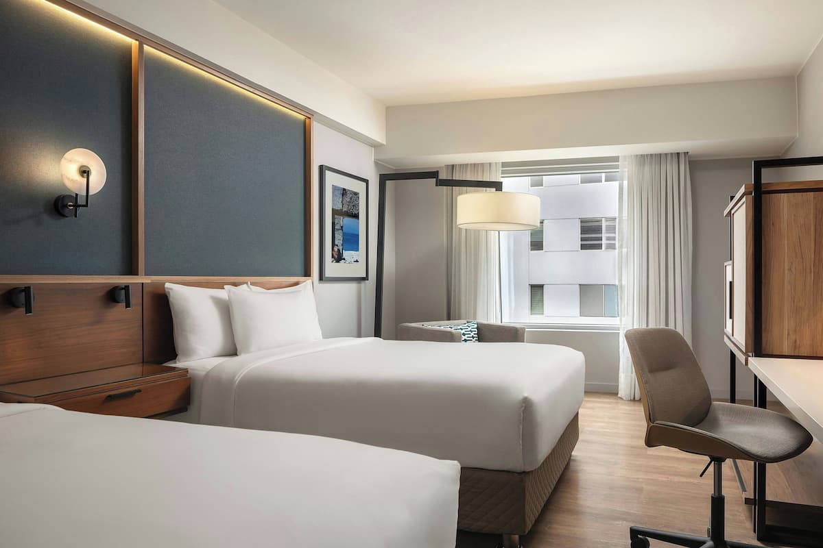 A hotel room with two beds, a desk, a chair, a lamp, a picture on the wall, and a window with curtains.