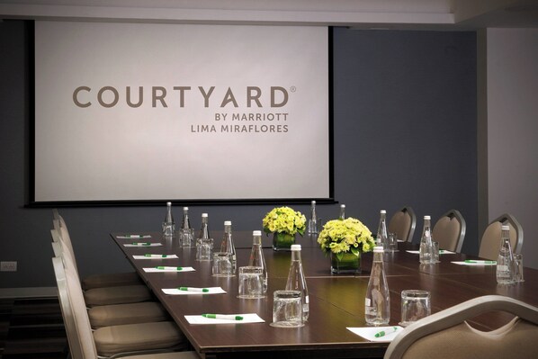 A conference room with a large screen displaying 'COURTYARD BY MARRIOTT LIMA MIRAFLORES'. The room has a long table with chairs, water bottles, and vases of yellow flowers.