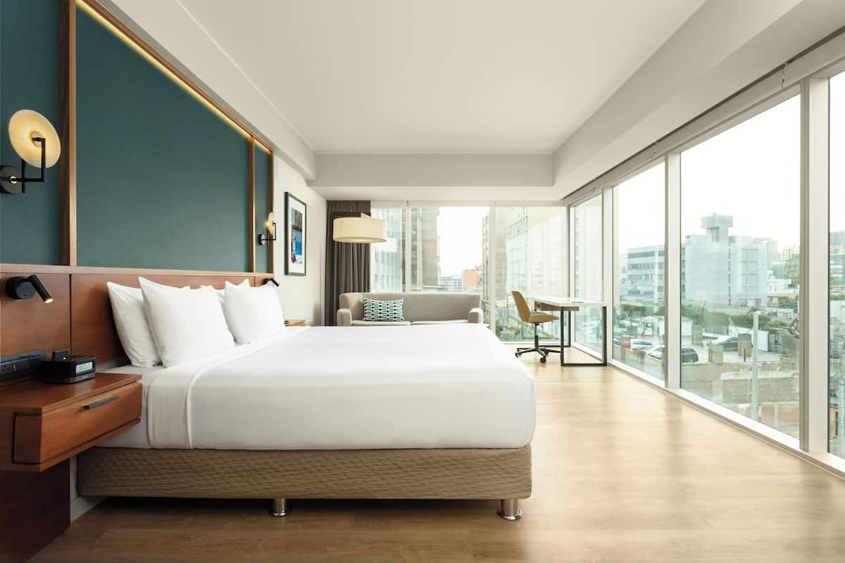 A modern hotel room with a large bed, a wooden nightstand, a desk, and a view of the city through large windows.