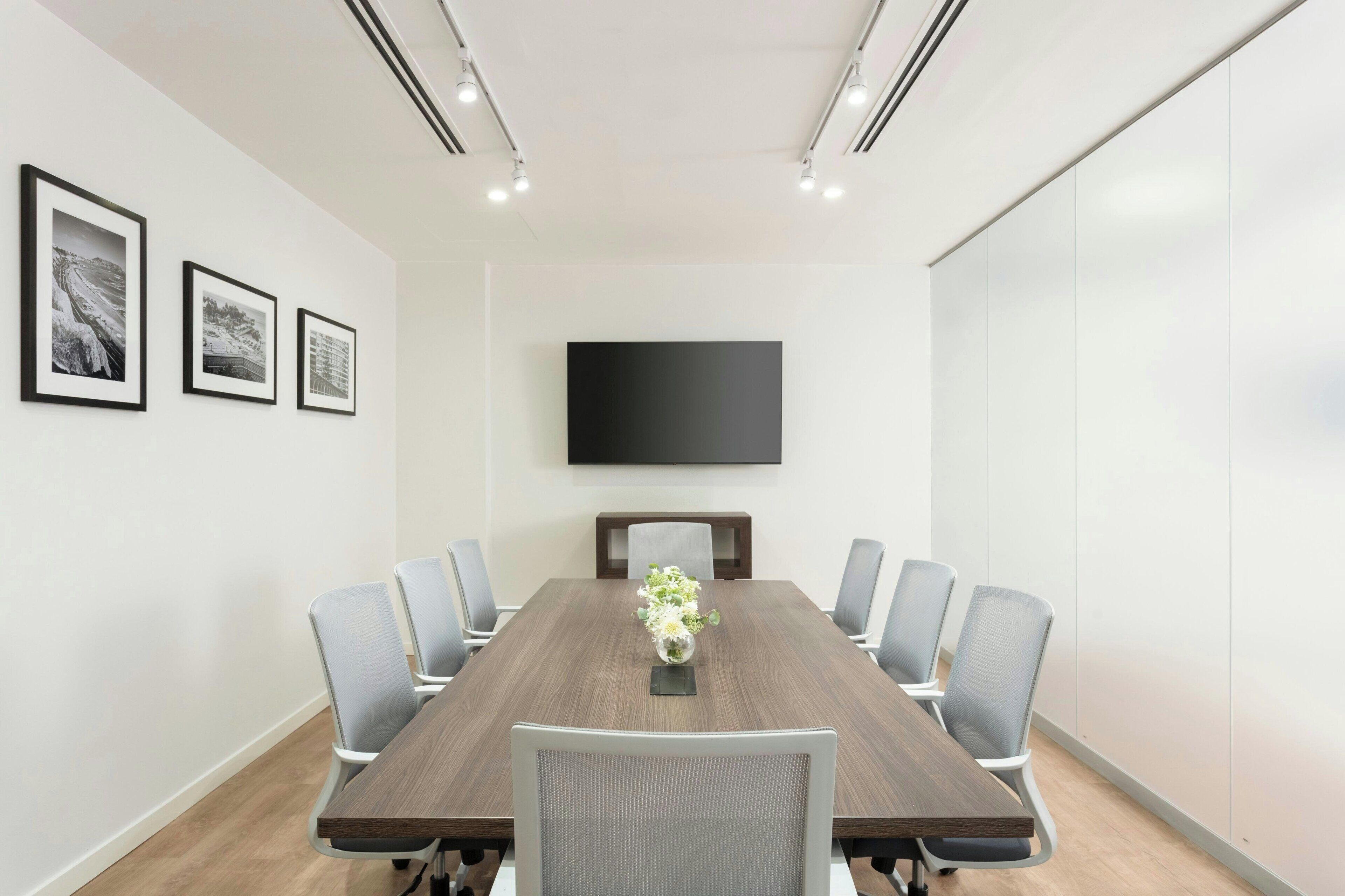 A conference room with a long table, six chairs, a flat-screen TV mounted on the wall, and three framed pictures on the wall.