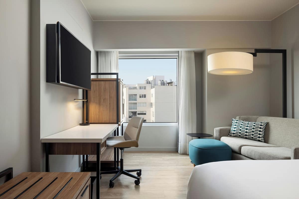 A hotel room with a desk, chair, sofa, ottoman, and a TV mounted on the wall.