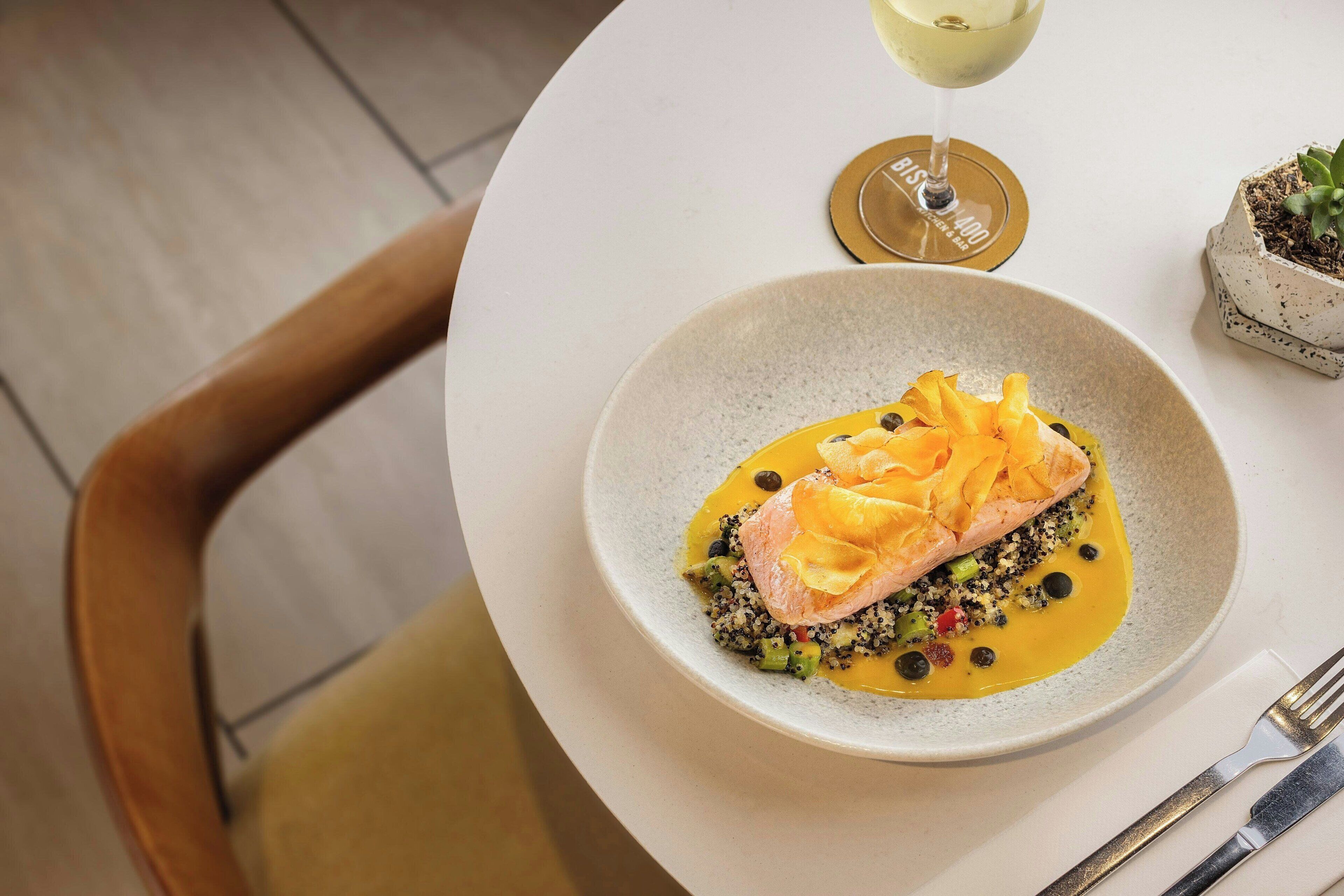 Una mesa de comedor con un plato de comida, una copa de vino blanco y una pequeña planta en maceta.