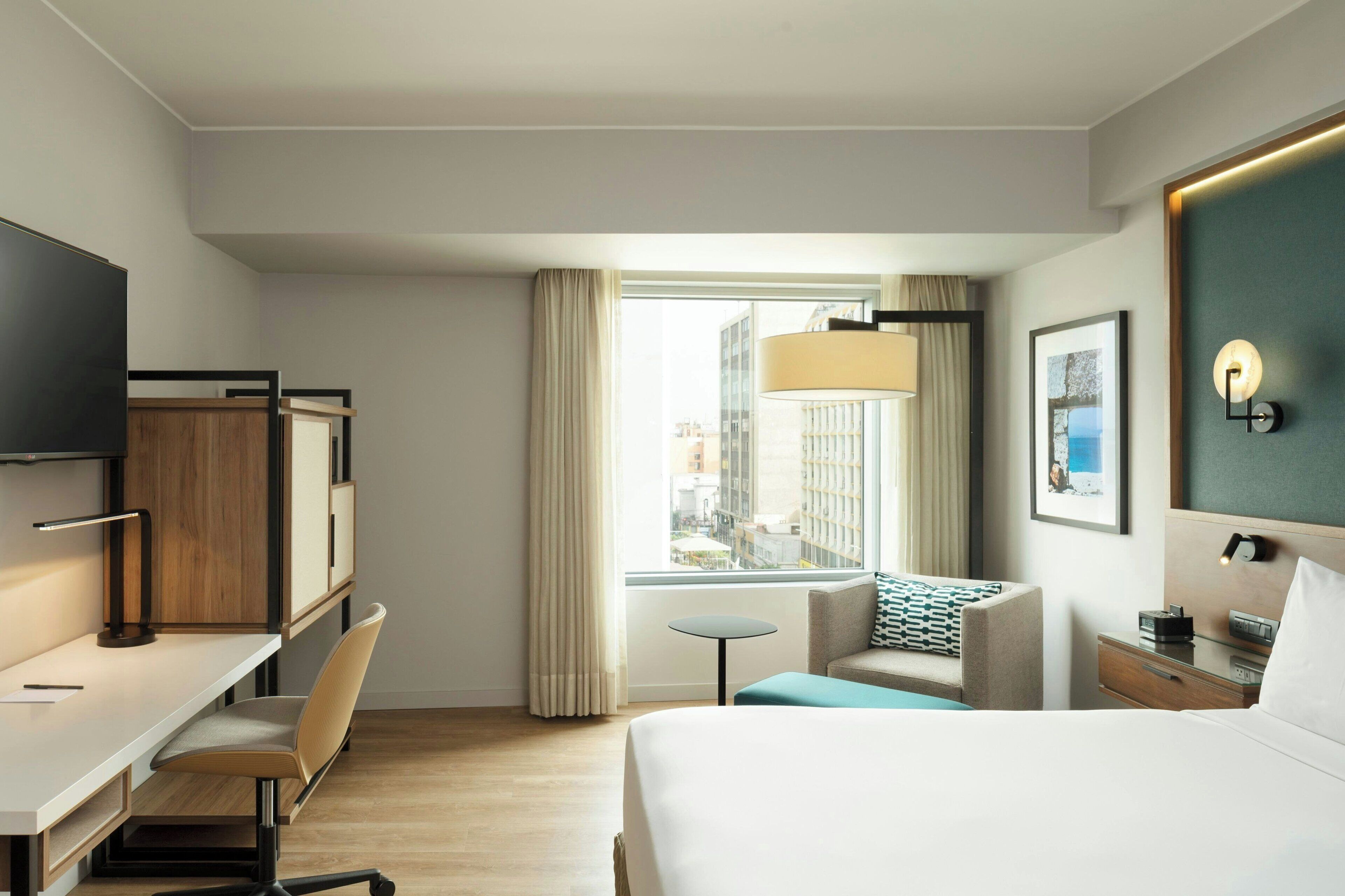 Camera d'albergo con un letto, una scrivania, una sedia e un tavolino. Si vede una finestra con tende, una lampada e un quadro appeso al muro.