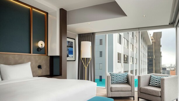 A modern hotel room with a large bed, two armchairs, a floor lamp, and a large window offering a city view. - Courtyard by Marriott Lima Miraflores (Lima)
