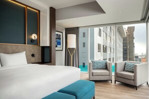 A modern hotel room with a large bed, two armchairs, a floor lamp, and a large window offering a city view.