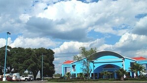 Un edificio grande de color azul con techo rojo, probablemente un hotel, rodeado de árboles y vehículos estacionados.