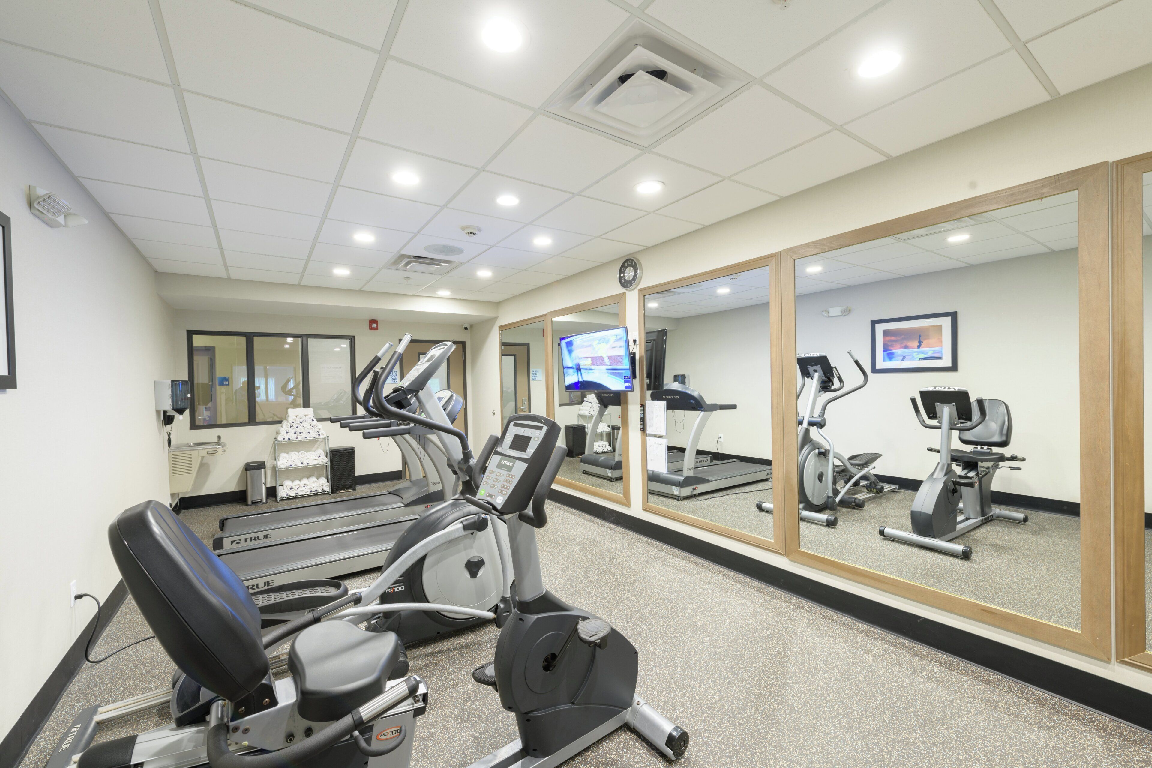 Ein Fitnessraum mit Trainingsgeräten, einer Spiegelwand und einer Uhr an der Decke.