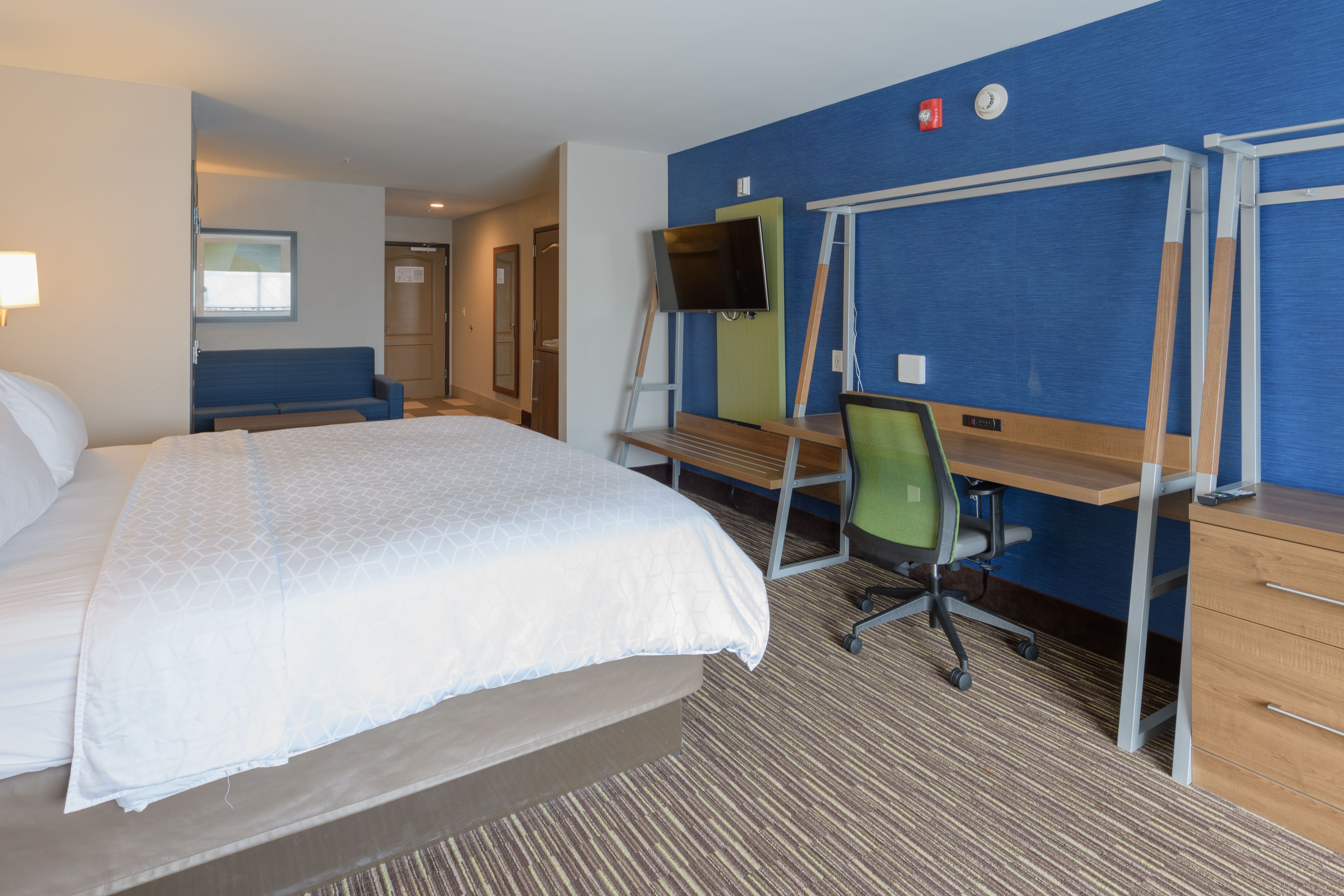 A hotel room with a large bed, a desk with a chair, a TV, and a blue accent wall.