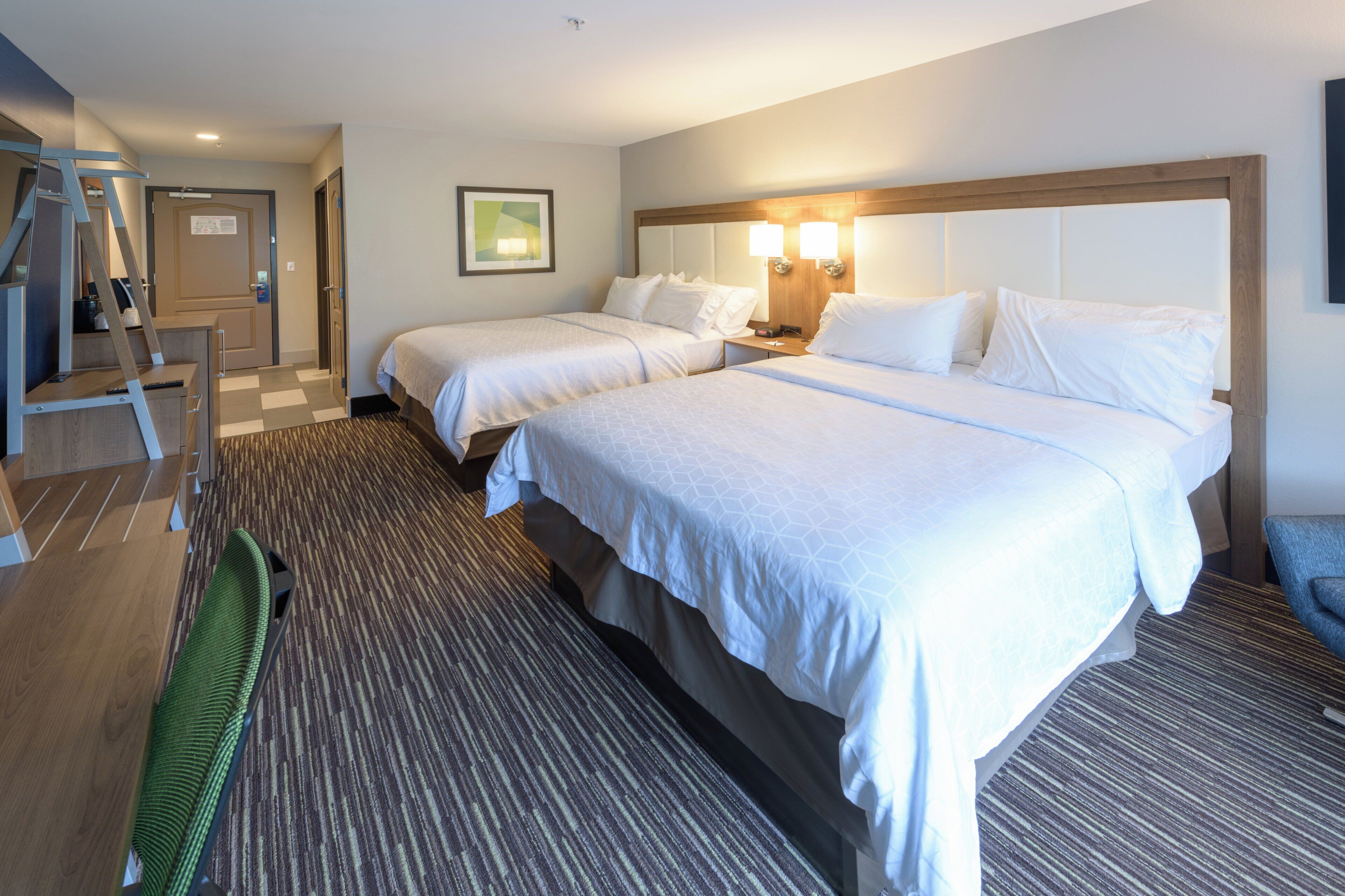 Ett hotellrum med två sängar, ett skrivbord, en stol, en stege och ett golv täckt av matta.