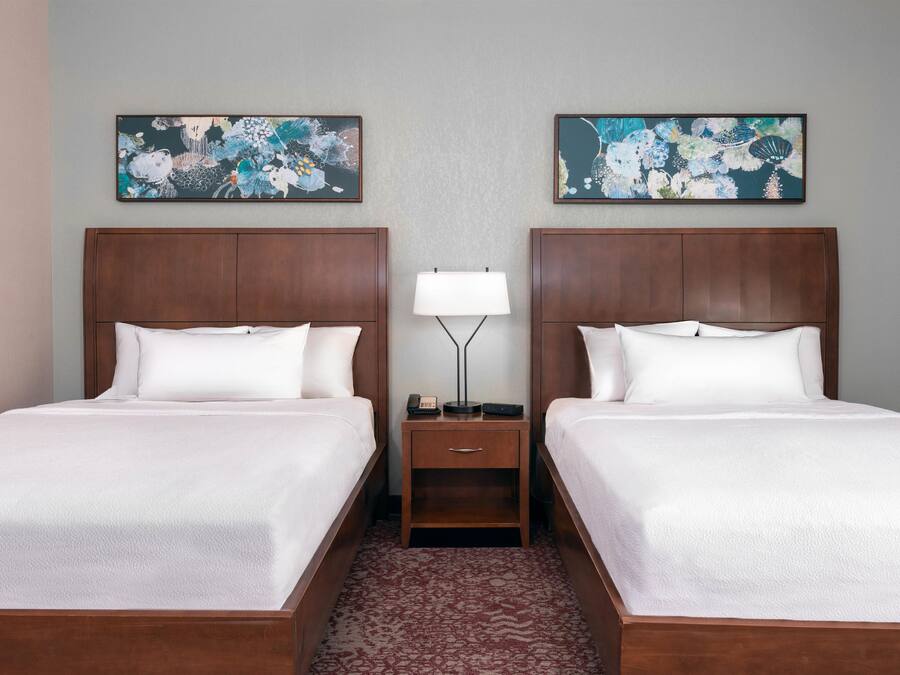 A hotel room with two beds, a nightstand with a lamp, and floral artwork on the walls.