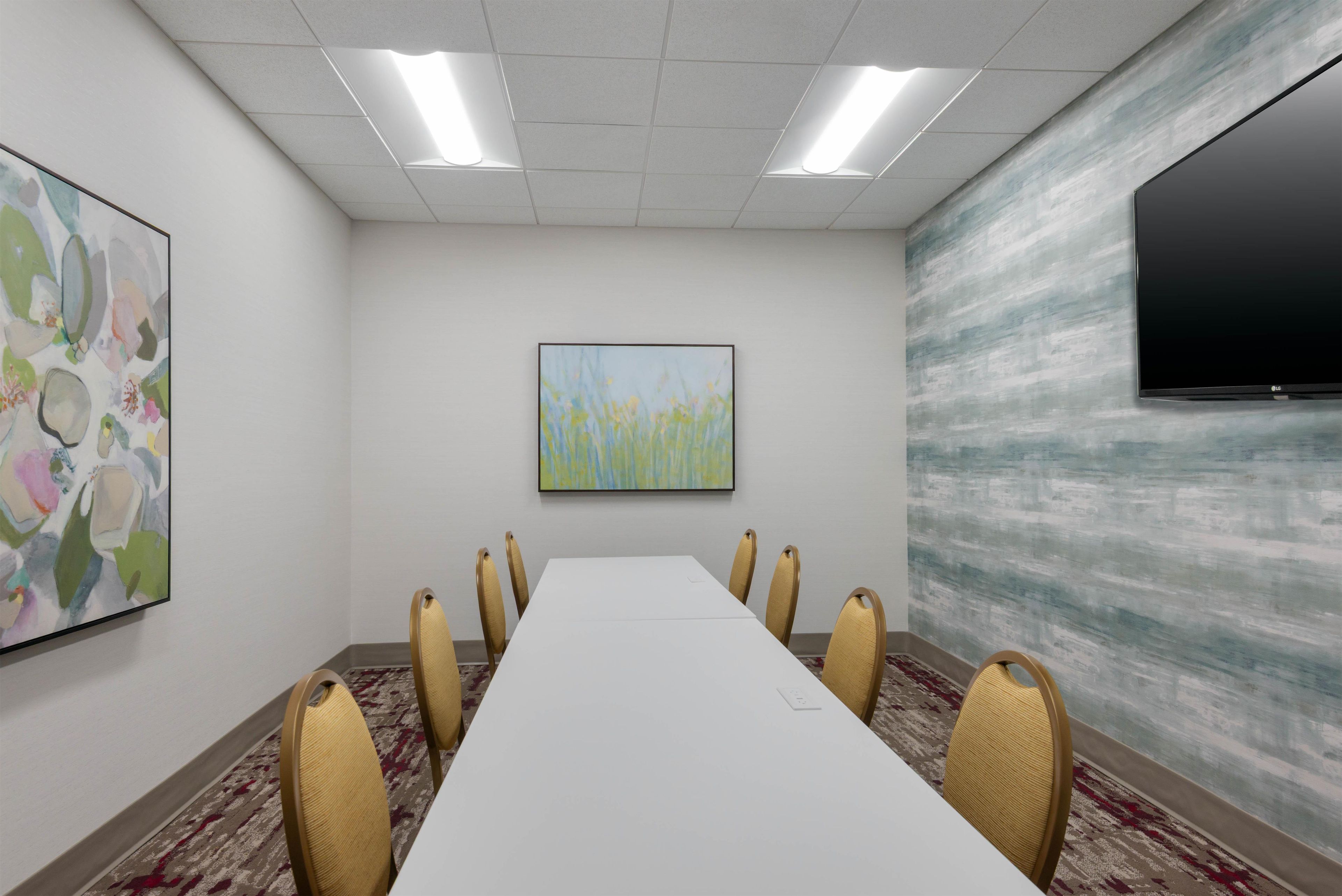 Uma sala de conferências com uma mesa comprida, seis cadeiras, um quadro grande, uma televisão e uma parede texturizada.
