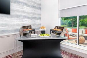 Una mesa de buffet con opciones de comida caliente y fría, un televisor de pantalla plana montado en la pared, y vistas a un jardín a través de grandes ventanales.