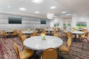 Un salón de conferencias con mesas redondas, sillas, un televisor de pantalla plana grande y vistas a zonas verdes por las ventanas.