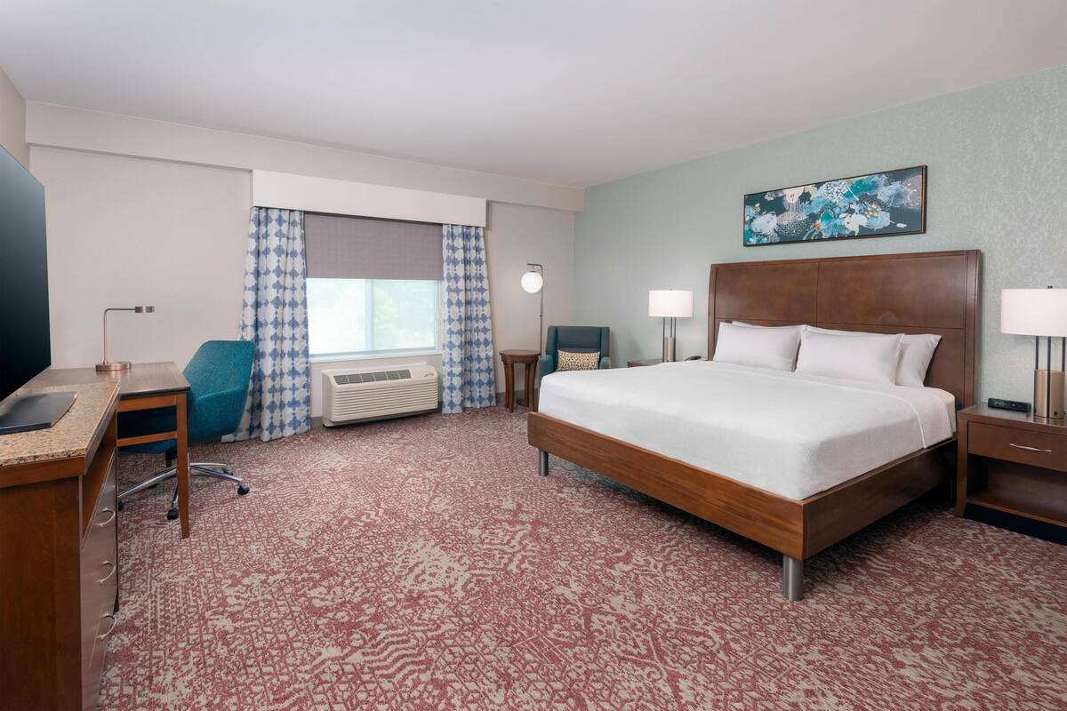 Habitación de hotel con cama, escritorio, silla, equipo de aire acondicionado y ventana con cortinas.