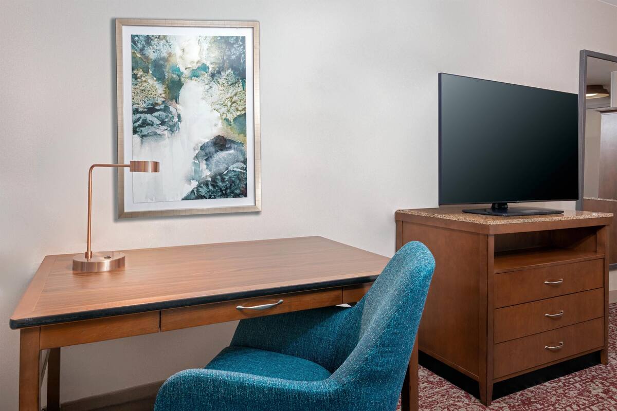 Una habitación con un escritorio de madera, una silla azul, un televisor de pantalla plana y un cuadro en la pared.