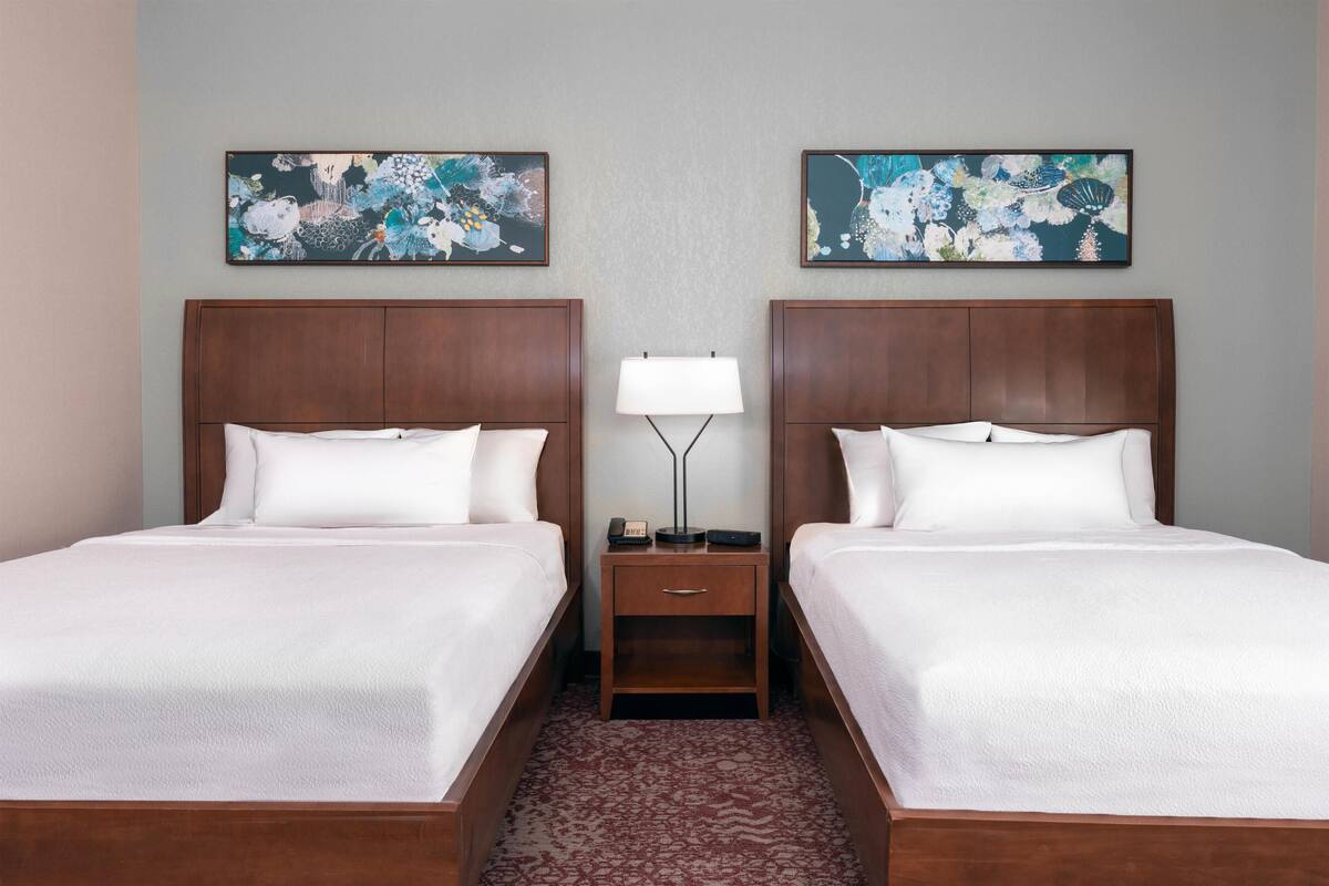 Habitación de hotel con dos camas, una mesita de noche con lámpara y cuadros con motivos florales en las paredes.