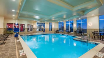 An indoor swimming pool with clear water, surrounded by lounge chairs and tables, and large windows offering views.