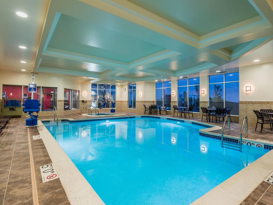 An indoor swimming pool with clear water, surrounded by lounge chairs and tables, and large windows offering views.