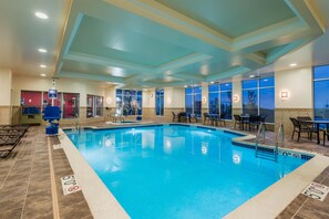 An indoor swimming pool with clear water, surrounded by lounge chairs and tables, and large windows offering views. - Hilton Garden Inn Wayne (Wayne)