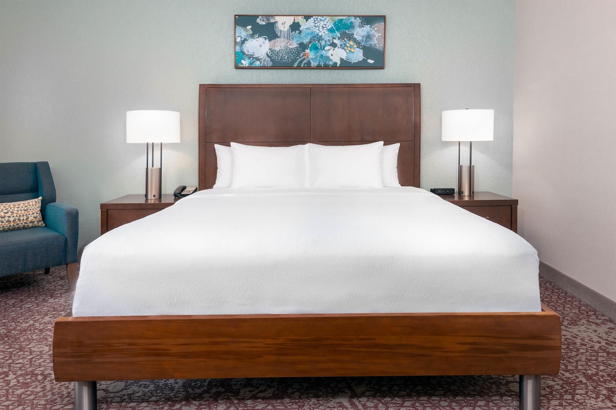 A hotel room with a large bed, two bedside tables, a nightstand, a blue armchair, and a painting on the wall.