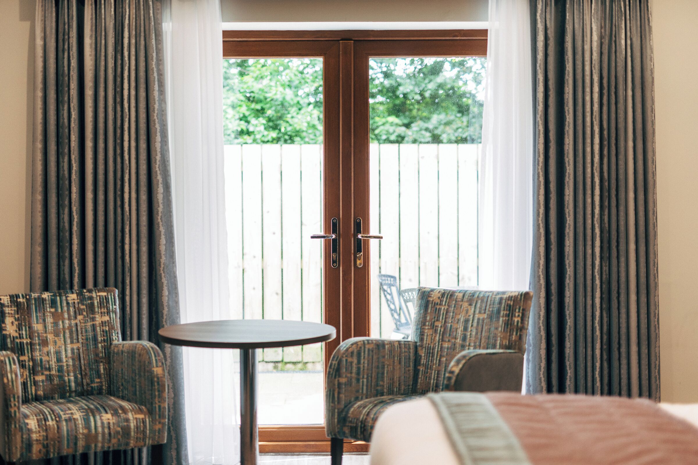 A room with a bed, two upholstered chairs, a small round table, and a sliding glass door leading to an outdoor area with a wooden fence and greenery.