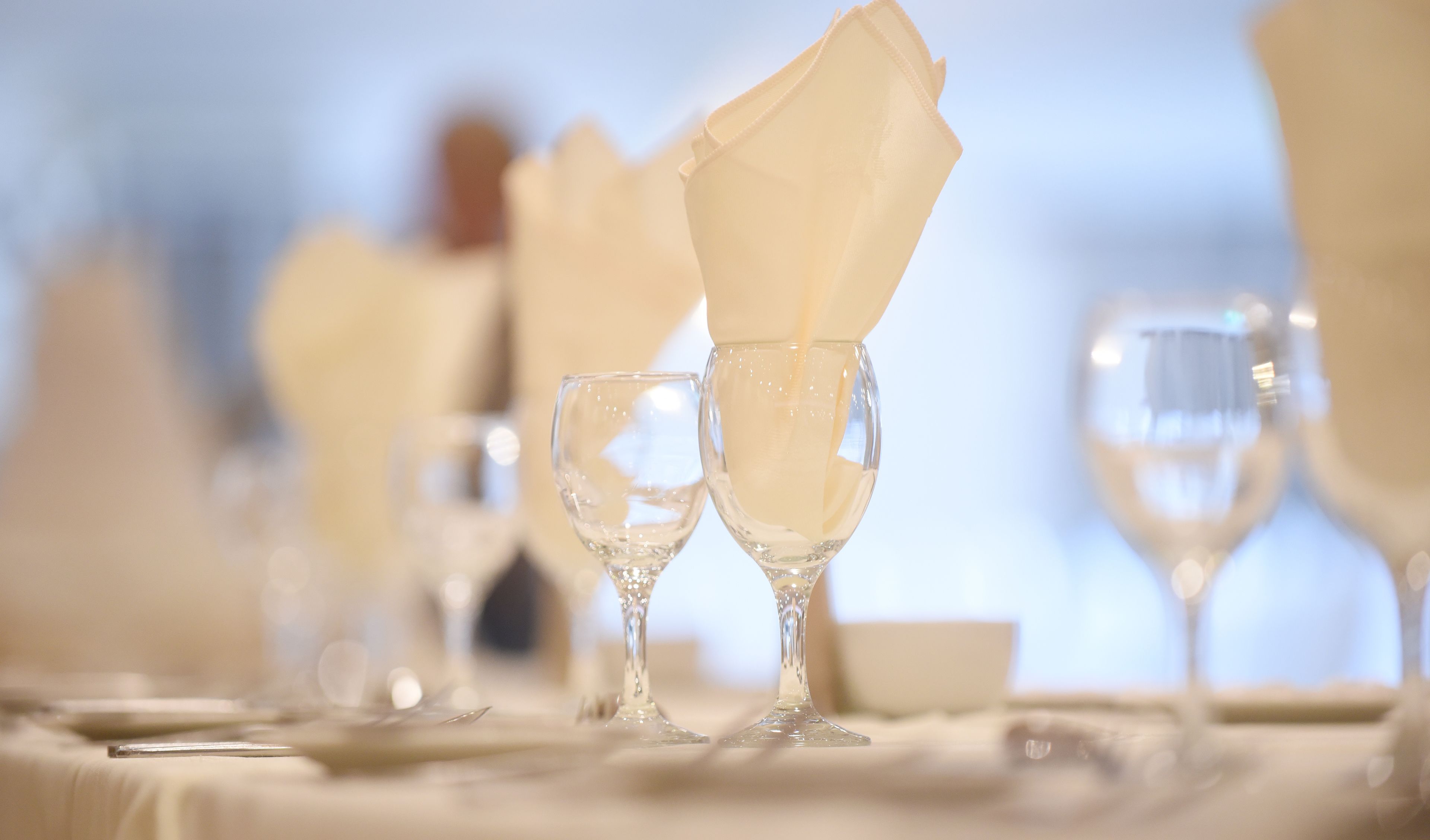 A table set with white napkins folded into a fan shape, clear wine glasses, and a blurred background suggesting a formal dining setting.